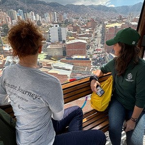 Two people look out a window over a dense population center with mountains in the background.
