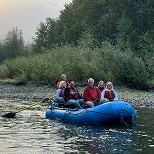A group of people sitting on a blue raft on a calm river with lush forest in the background.