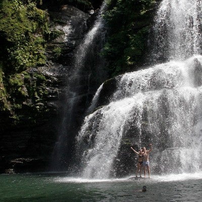 Two people stand at the base of a large waterfall in a tropical rainforest