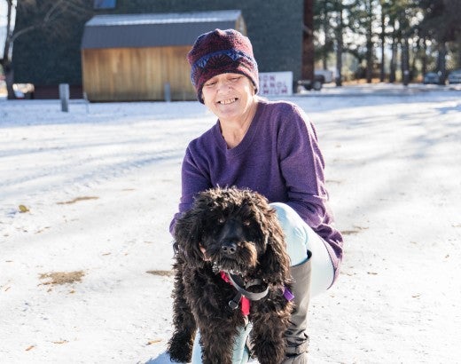 A woman in a purple beanie and shirt holds a black, curly haired dog in the snow
